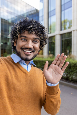 Smiling businessman in casual attire waving at camera during video call using phone. Image captures friendly, cheerful mood in urban business setting, with modern office in background.の写真素材