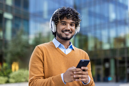 Confident young man with curly hair wearing headphones and holding smartphone outside contemporary glass office building. He looks happy and relaxed, dressed in casual sweaterの写真素材