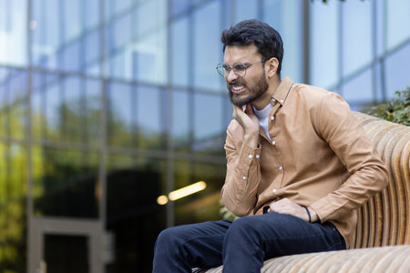 Man shows signs of toothache, seated on outdoor bench, holding jaw with pained expression. Casual clothing and glasses suggest relaxed setting. Image captures moment of discomfortの写真素材