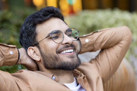 Smiling young man in casual wear relaxes outdoors. His joyful expression and comfortable posture convey feelings of happiness and leisure. Ideal image for concepts of relaxation, positivityの写真素材