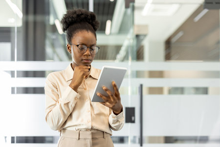 African American businesswoman in office examines tablet thoughtfully. Focus on technology and innovation highlights professional dedication. Modern setting and attire emphasize commitment to workの写真素材