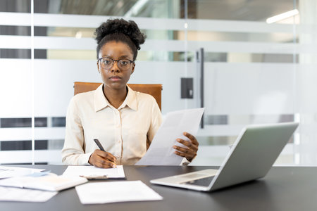 African American businesswoman in office with laptop holding documents while writing. Professional environment emphasizes focus, analysis, and productivity in workplace setting.の写真素材