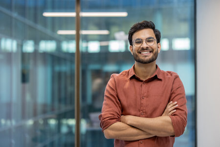 Smiling young professional standing with arms crossed in modern office. Confident individual exuding positivity and success. Represents business spirit, leadership, and motivation within workplaceの写真素材