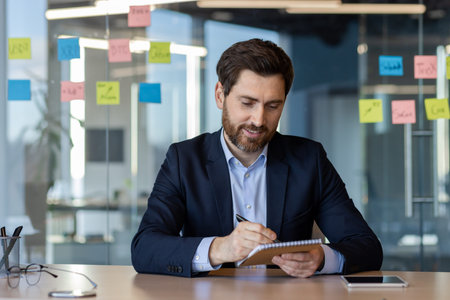 Businessman in modern office writing notes with focused expression. Visible sticky notes on glass...