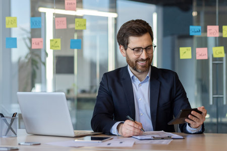 Smiling businessman in glasses working at desk with laptop, phone, and documents. Holding calculator, checking data. Bright modern office setting with financial notes.の写真素材