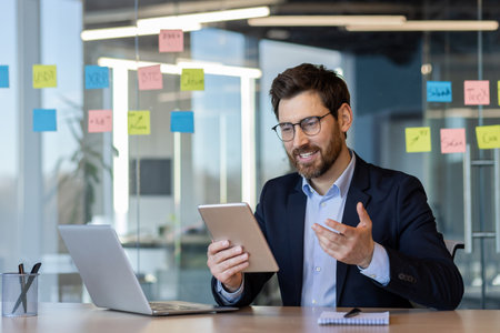 Confident businessman in office conducting video call on tablet. Engaged in virtual communication, conveying ideas effectively. Professional setting emphasizes technology and modern work environment.の写真素材