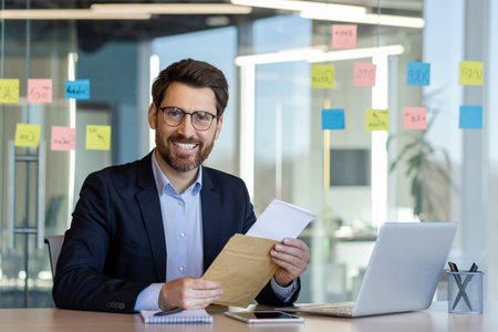 Smiling businessman holding document at office desk with laptop and notepad. Professional environment suggesting focus and productivity.の写真素材