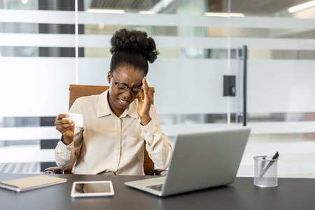 Young businesswoman experiencing headache while working on project. She holds tissue and uses laptop at office desk, conveying stress and effort in achieving professional tasks.の写真素材