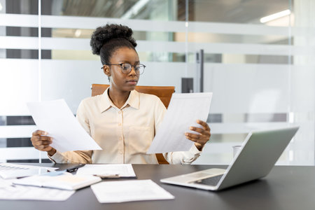 African American businesswoman in office setting reviewing documents with focused expression....
