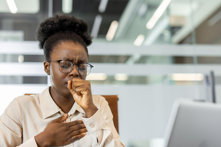 African American businesswoman experiencing discomfort and coughing in office. Depicts stress, illness or fatigue in workspace. Highlights challenges faced by professionals managing healthの写真素材