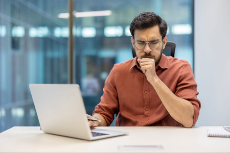 Male office worker sits at desk wearing glasses looking concerned, experiencing cough symptoms while...