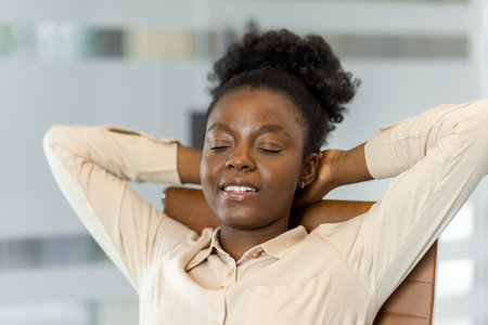 African American businesswoman relaxing in office chair with closed eyes, enjoying peaceful moment during workday. Professional attire, calm expression reflecting stress relief and workplace balanceの写真素材