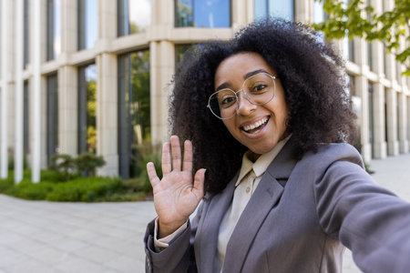 Confident African American business woman waving during phone video call. Standing in front of contemporary office building. Professional and friendly demeanor enhances business communication.の写真素材