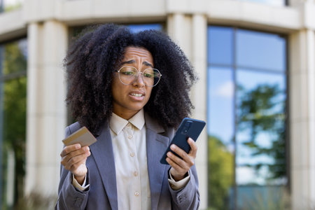 African American businesswoman expressing concern holding credit card and smartphone outside modern office building. Represents online banking, financial management, digital transactions stress.の写真素材