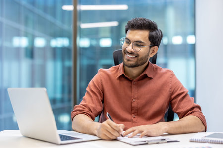 Businessman in glasses smiling while working on laptop and writing notes. Depicts professional...