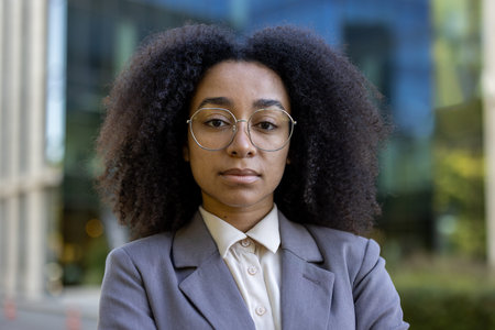 Confident African American business woman with curly hair and glasses standing outdoors, showcasing...