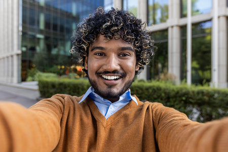 Smiling young man taking selfie outside modern office building, wearing casual attire. Capturing...