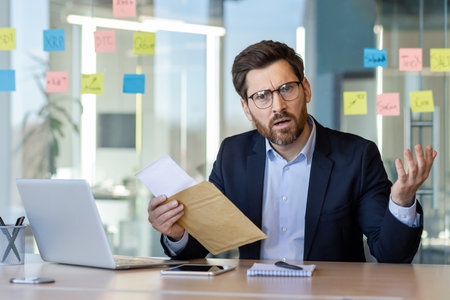 Businessman sitting at office desk holding envelope and gesturing with confusion. Frustrated expression suggests receiving unexpected news.の写真素材