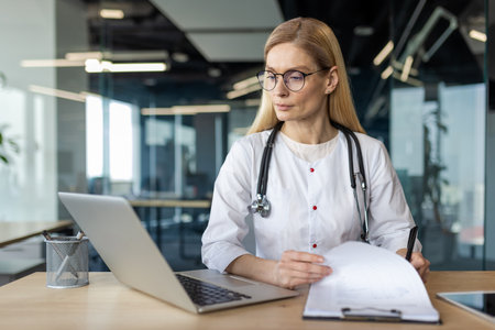 Professional female doctor in white coat with stethoscope working on laptop and examining medical documents. Scene conveys focus, expertise, and concentration in healthcare and medical field.の写真素材