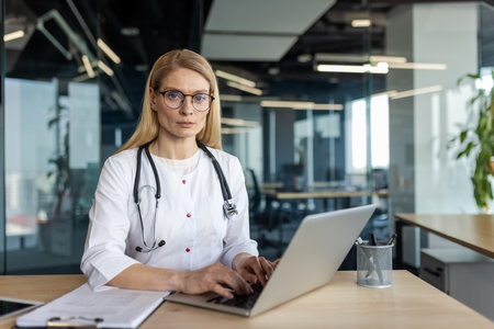 Professional female doctor wearing glasses confidently working on laptop in modern office setting. Focused on medical tasks with stethoscope and documents nearbyの写真素材