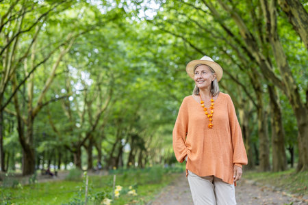 Smiling elderly woman walking in park, wearing a straw hat and orange sweater. Image captures joy, relaxation, and connection with nature, highlighting peaceful lifestyle and positive attitude.の写真素材