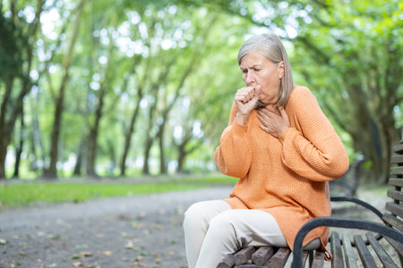 Senior woman sitting on park bench struggles with breathing. Image captures concern and health issues in tranquil natural environment. Depicts aging, respiratory problems, need for medical attention.の写真素材