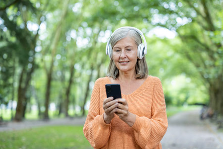 Senior woman wearing headphones, using phone outdoors in park setting. She appears happy, engaged...