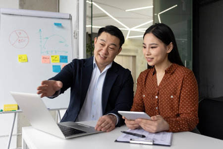 Asian business professionals engaged in collaborative work at office desk with laptop and tablet. Man explaining concepts using digital tools. Whiteboard with graphs and notes in background.の写真素材