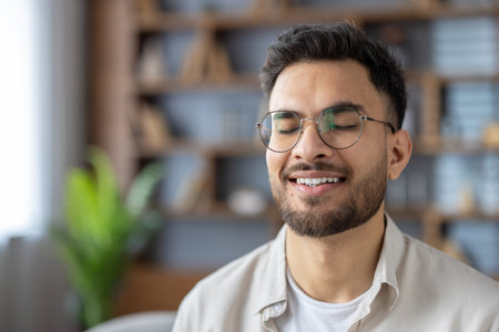 Young man with glasses smiling peacefully while meditating indoors. Capturing moment of relaxation, serenity, and mindfulness. Closed eyes convey sense of calm and inner peace tranquil environment.の写真素材