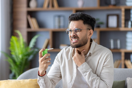 Sick young Indian man sitting on sofa at home, holding hand to neck and using medical spray for sore throat.の写真素材