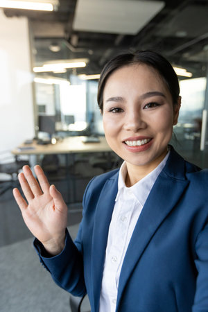 Asian business woman smiling and waving during video call in office. Professional attire and friendly demeanor highlight modern communication. Ideal for themes of business, technology, and networkingの写真素材