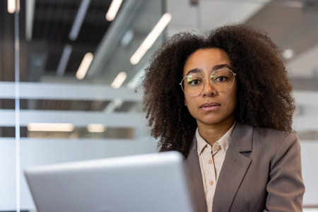 Close-up portrait of a young African-American woman in a business suit sitting in an office at a desk in front of a laptop, looking seriously at the camera.の写真素材