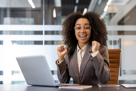 Portrait of a happy African-American woman in the office, sitting in a suit at the desk, rejoicing in success, raising her hands up and smiling at the camera.の写真素材
