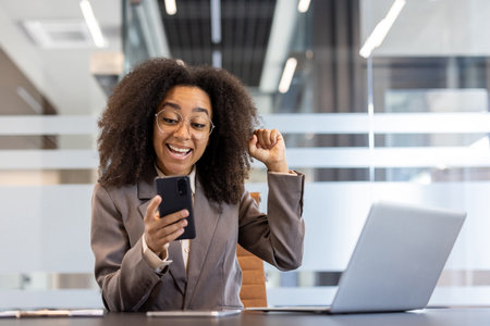 African American young woman in a business suit sitting in the office at the desk, looking at the phone and happy with success raising her hand up.の写真素材