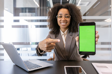 A young African American woman in a suit and glasses is sitting in the office at a desk with a laptop and pointing her finger at a green blank screen of a phone, looking and smiling at the camera.の写真素材