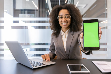 Close-up portrait of a smiling young African-American woman in a business suit and glasses sitting at a desk in an office, showing a mobile phone with a mock-up green screen to the camera.の写真素材