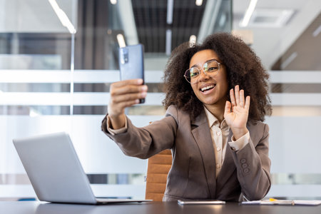 Happy and smiling young African American woman in a suit sitting at the desk in the office, talking on a video call on the phone and waving at the camera.の写真素材