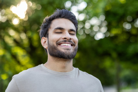 Portrait young man with beard standing outdoors in park. He smiles with eyes closed, capturing peaceful moment. Expression conveys happiness, serenity, and contentment amidst natural surroundings.の写真素材