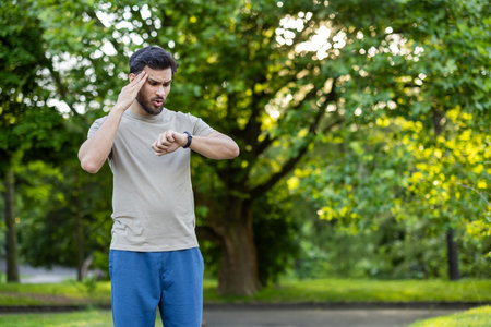 Young male athlete outdoors checking smartwatch. Man appears tired after training session while standing in sunlit park. Concept of fitness, exercise, wearable technology.の写真素材