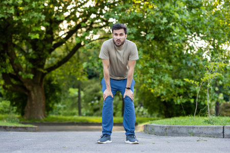 Young sportsman resting after an intense exercise session in park. Dressed in casual sportswear, he leans forward catching breath, surrounded by serene green environment.の写真素材