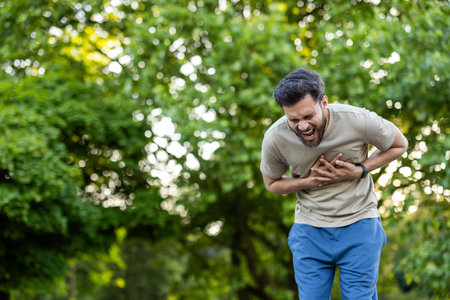 Man suffers from sudden chest pain exercising in park. He appears distressed, clutching his chest in...