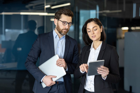 Business professionals engaged in discussion using tablet in office setting. Man holding laptop while woman demonstrates with tablet, showcasing teamwork and communication in corporate environmentの写真素材