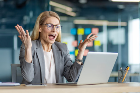 Joyful businesswoman expressing excitement at office desk with laptop. Professional wearingsuit celebrating success. Positive emotions and workplace achievement in modern business setting.の写真素材