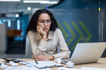 Office worker feeling dissatisfied with report results. Person sits at desk with expressions of upset while reviewing disappointing paperwork on laptop. Surrounding desk cluttered with crumpled paperの写真素材