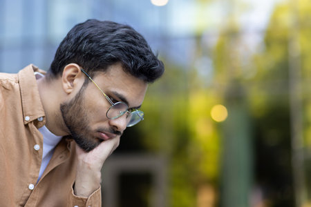 Pensive man wearing glasses and brown shirt looks down thoughtfully, conveying contemplation and focus. Urban setting in background enhances mood of introspection and focus.の写真素材