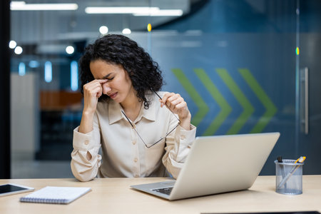 Stressed female office worker rubbing eyes in front of laptop indicating workplace pressure. Notebook on desk suggest busy environment. Image conveys stress, fatigue, and need for a break.の写真素材