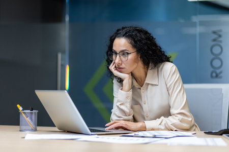 Professional businesswoman with glasses using laptop in office. Focused expression, analyzing documents on desk. Office setting suggests roles like manager, analyst, or consultant.の写真素材