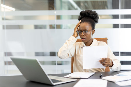 Young African American woman at workplace feeling stressed while working in office. Holding documents, looking at laptop with hand on forehead, experiencing workplace stress, frustration during workの写真素材