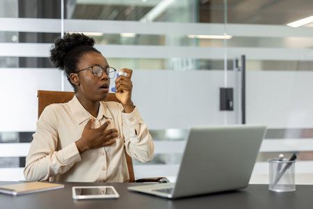 Young african american woman experiencing asthma attack in office environment uses inhaler while sitting at desk with laptop and phone nearby, demonstrating health challenges in workspaces.の写真素材