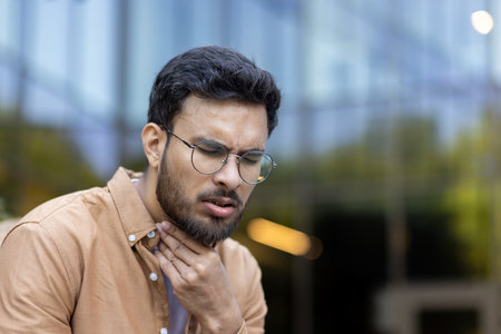 Portrait of man with beard and glasses holding throat indicating discomfort or sore throat. Outdoors in casual attire, expressing health concern, illness, allergy. Captures moment pain or irritation.の写真素材